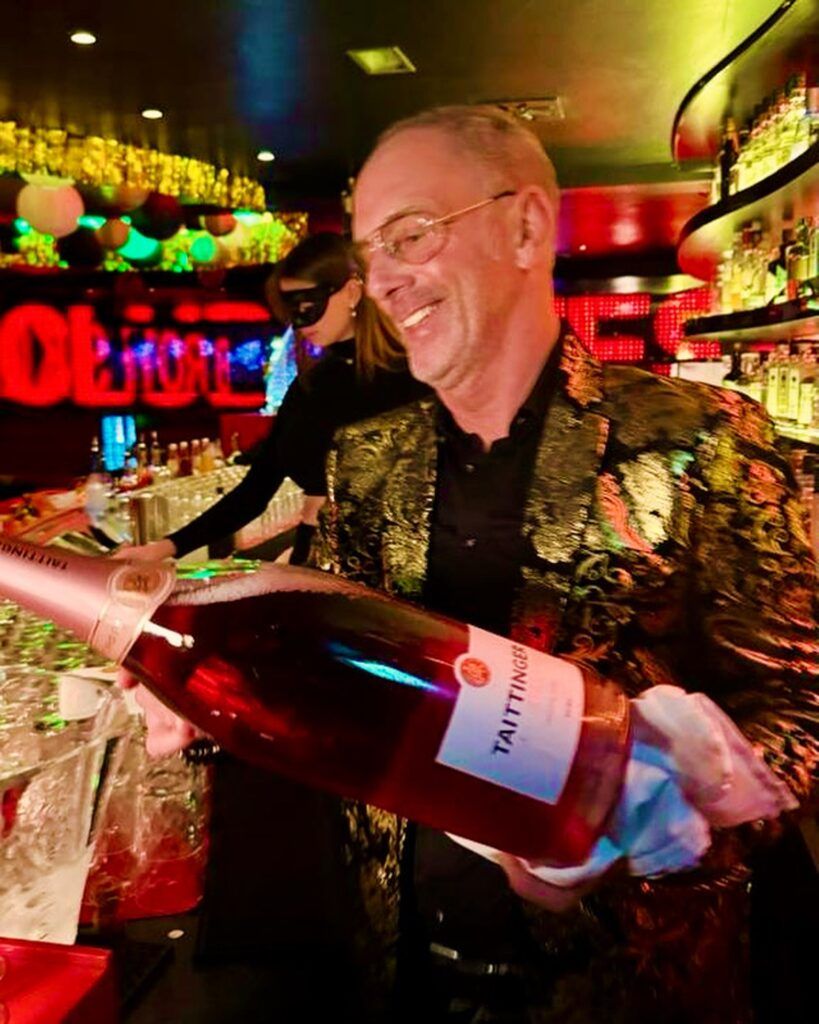 Ein lächelnder Mann in einer gemusterten Jacke hält eine große Flasche Champagner an einer Bar.