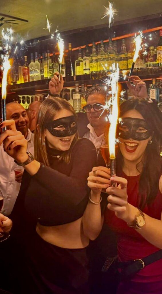 Gruppe maskierter Menschen feiert mit Wunderkerzen und Getränken auf einer Party.